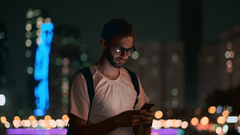 A-young-man-with-glasses-at-night-looks-at-the-smartphone-screen-and-writes-text-messages-for-his-blog-on-social-networks.-Reads-information-and-makes-a-trip-martour